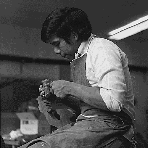 [Detail of] Cherokee high school student seated on a desk wearing a work apron carving a small animal statue.